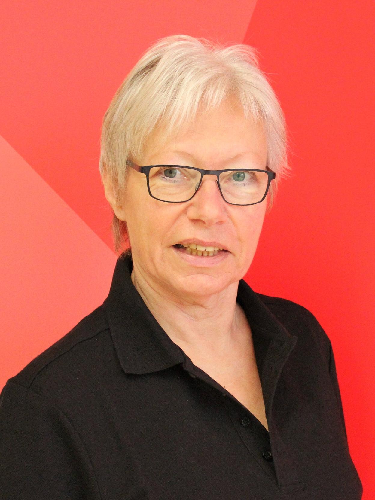 Portr&auml;t einer Frau mit kurzen grauen Haaren und Brille, im schwarzen Polohemd vor einem roten Hintergrund.