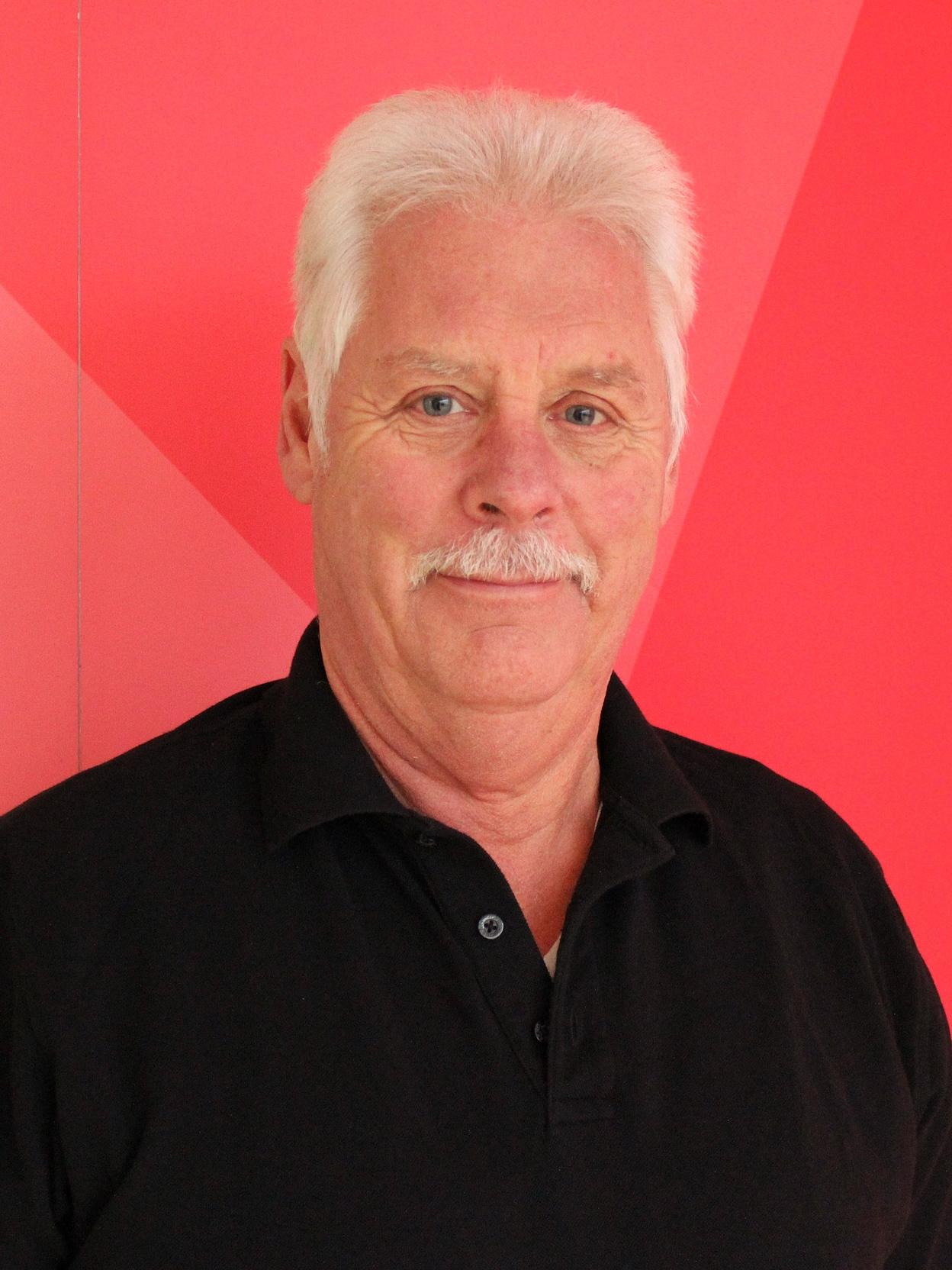 Portr&auml;t eines &auml;lteren Mannes mit hellem Haar und Schnurrbart, tr&auml;gt ein schwarzes Hemd vor rotem Hintergrund.