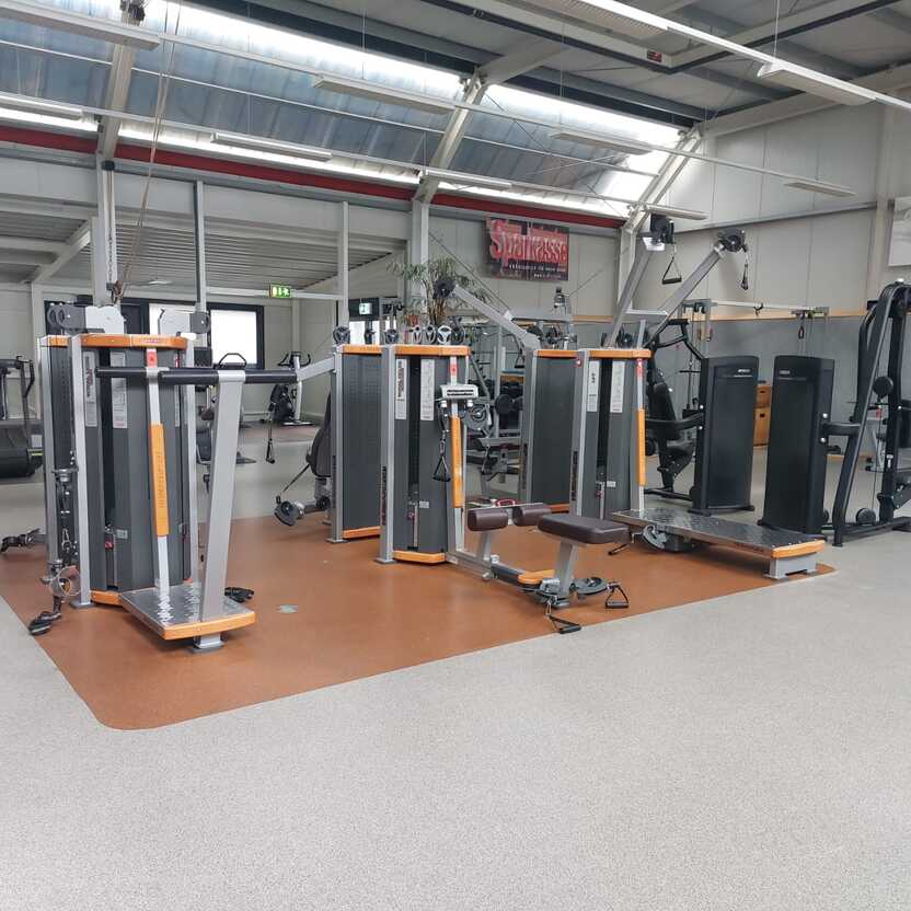 Fitnessstudio mit mehreren Trainingsger&auml;ten auf einem hellen, offenen Bodenbelag. Durch gro&szlig;e Fenster f&auml;llt Tageslicht ein.