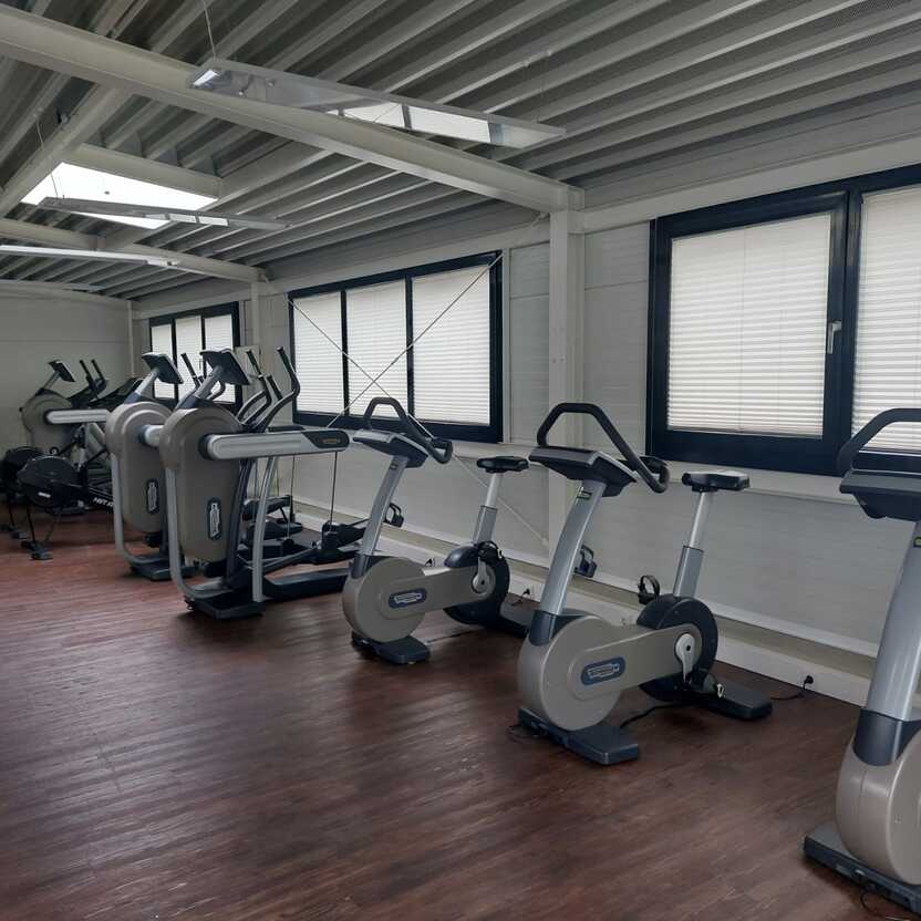 Fitnessraum mit mehreren modernen Ergometern und einem Holzfu&szlig;boden, beleuchtet durch Fenster mit Jalousien.