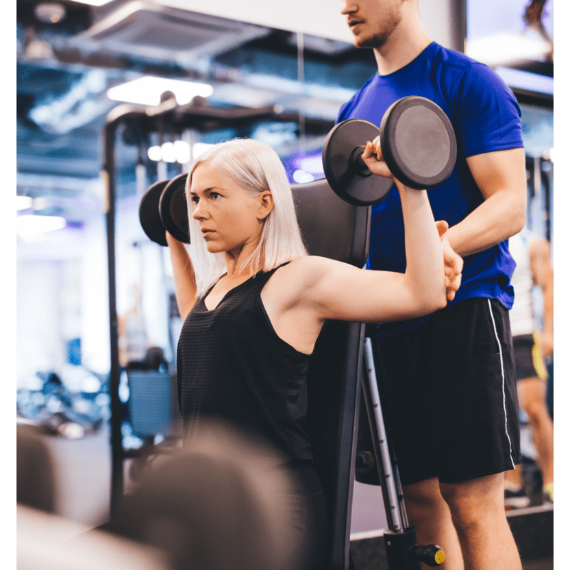 Frau mit blonden Haaren hebt Gewichte auf einer Schulterpresse, w&auml;hrend ein Trainer sie unterst&uuml;tzt.