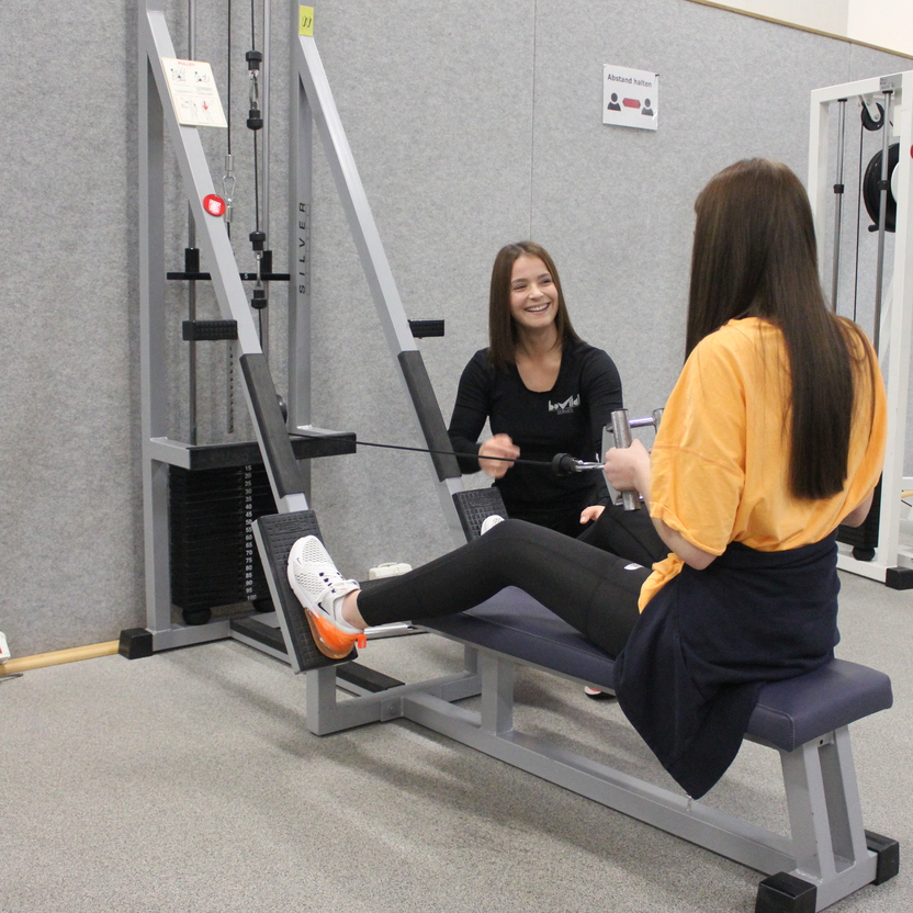 Zwei Frauen stehen in einem Fitnessstudio, eine sitzt an einem Kabelzugger&auml;t und l&auml;chelt, die andere gibt Anweisungen.