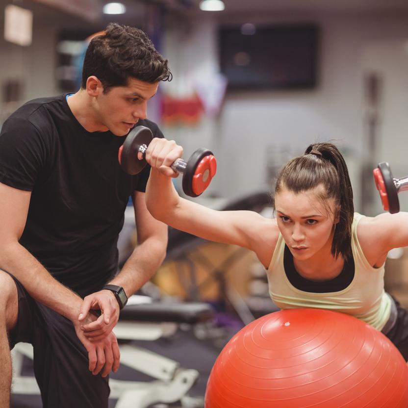 Eine Frau mit Hanteln &uuml;bt auf einem Gymnastikball, w&auml;hrend ein Mann sie beobachtet und anleitet.