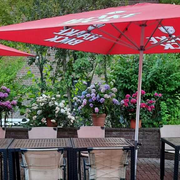 Terrasse mit mehreren Tischen, St&uuml;hlen und bunten Blumen, &uuml;berdacht mit roten Sonnenschirmen.