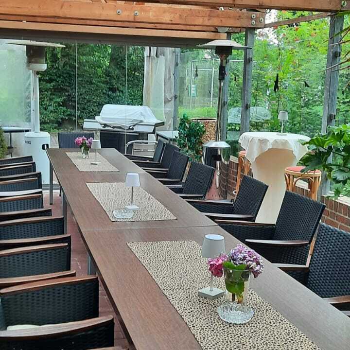 Gedeckte Terrasse mit einem langen Holztisch, St&uuml;hlen, Pflanzen und einem Grill im Hintergrund.