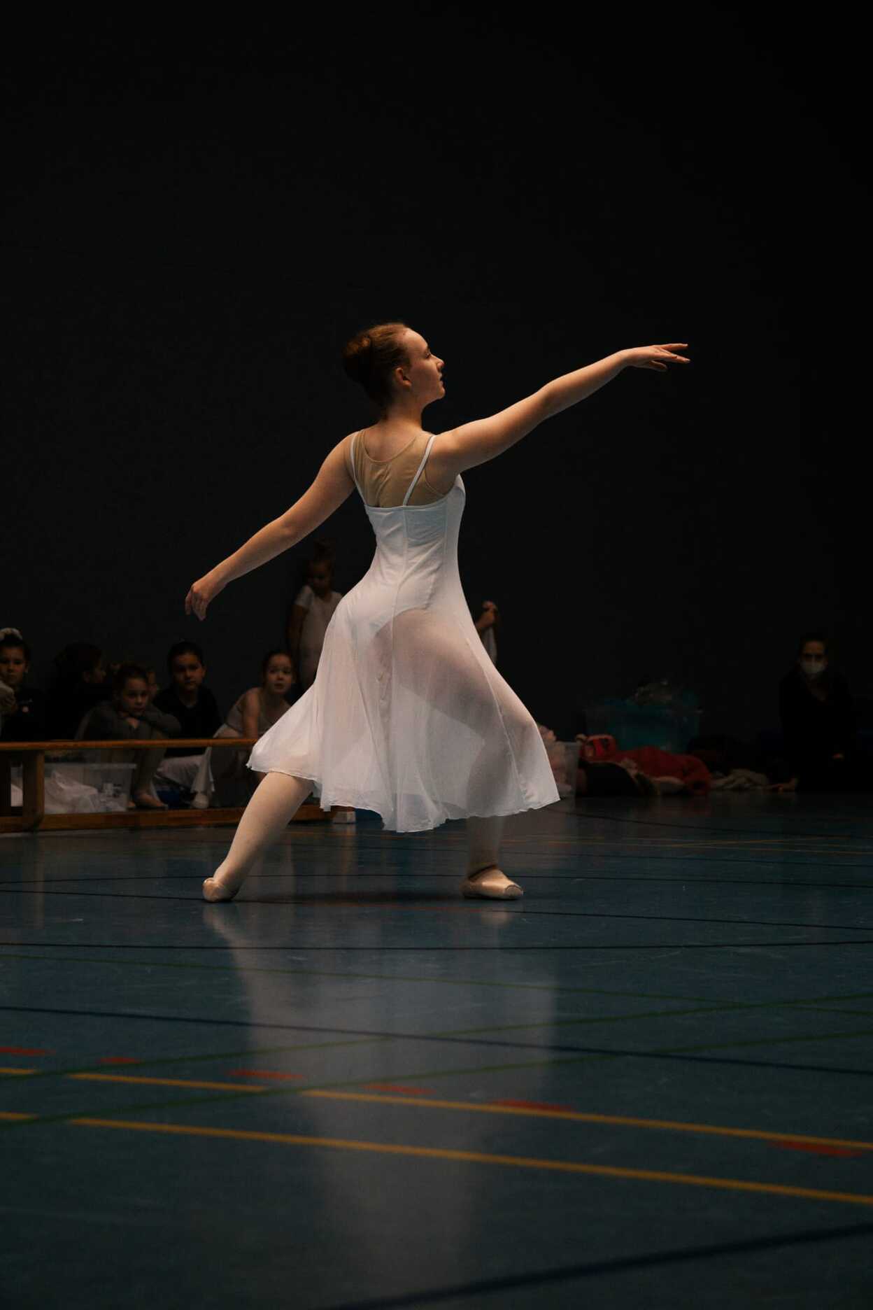 Ballettt&auml;nzerin in einem wei&szlig;en Kleid, die sich auf einer Sportfl&auml;che elegant dreht, Zuschauer im Hintergrund.