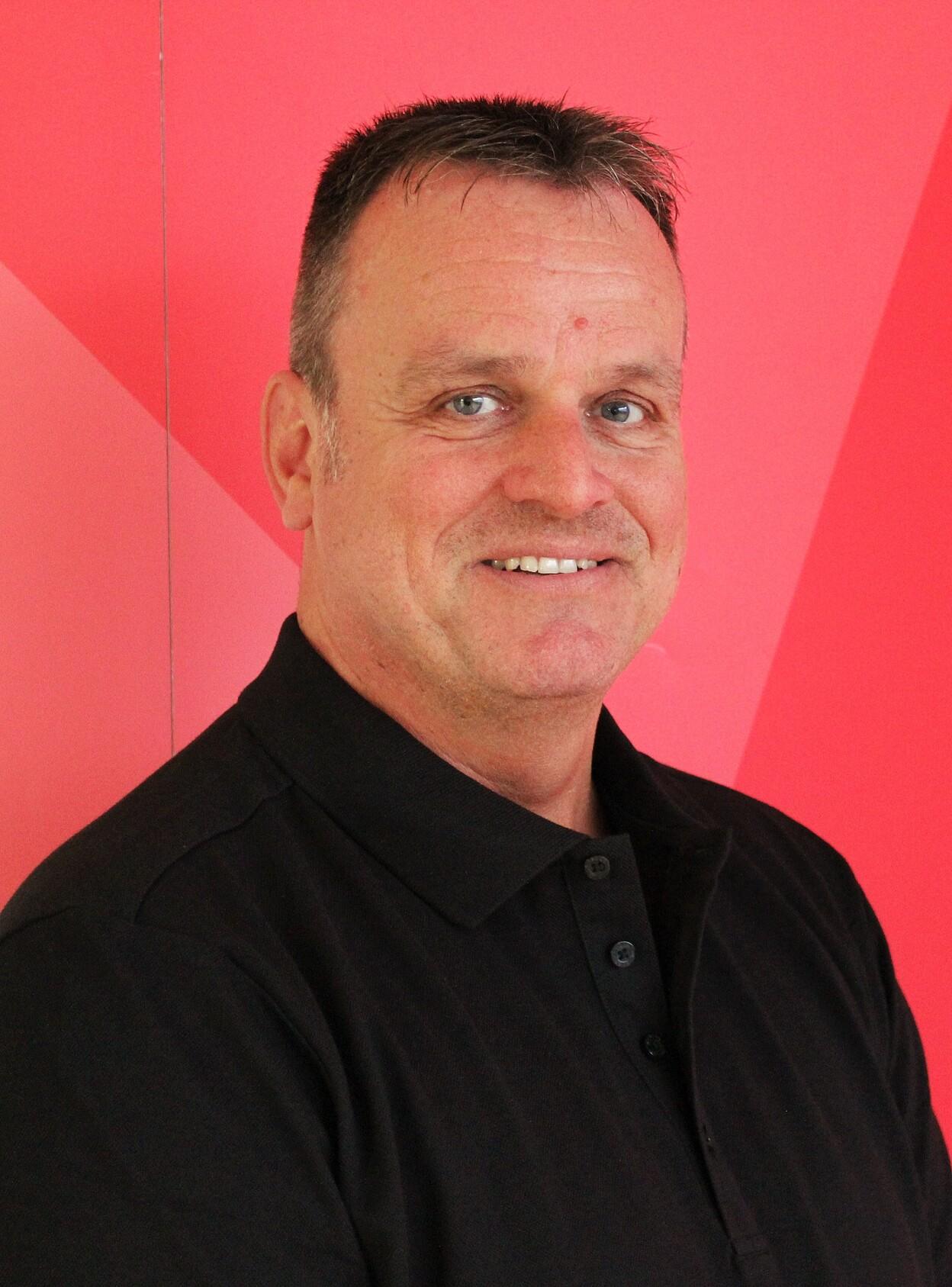 Portr&auml;t eines l&auml;chelnden Mannes mit kurzer, grauer Frisur vor einem pinken Hintergrund. Er tr&auml;gt ein schwarzes Poloshirt.