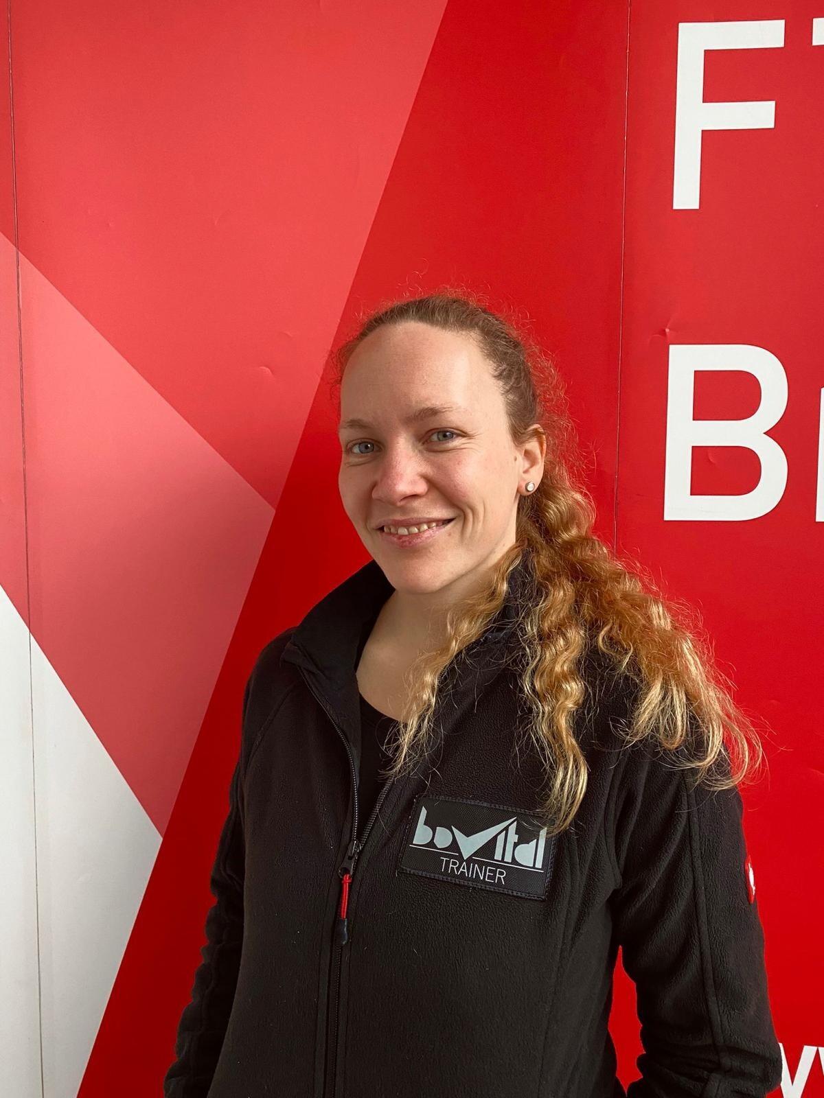 Frau mit langen, lockigen Haaren in schwarzer Trainerjacke steht vor rotem Hintergrund mit dem Schriftzug &bdquo;FITNESS&ldquo;.