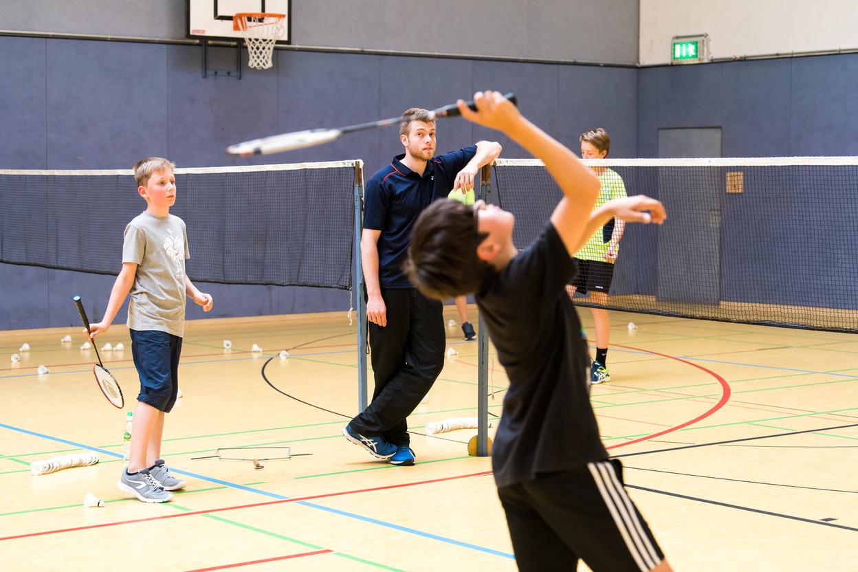 Vier Kinder spielen Badminton in einer Sporthalle, w&auml;hrend ein Trainer an der Seite steht und die &Uuml;bungen beobachtet.