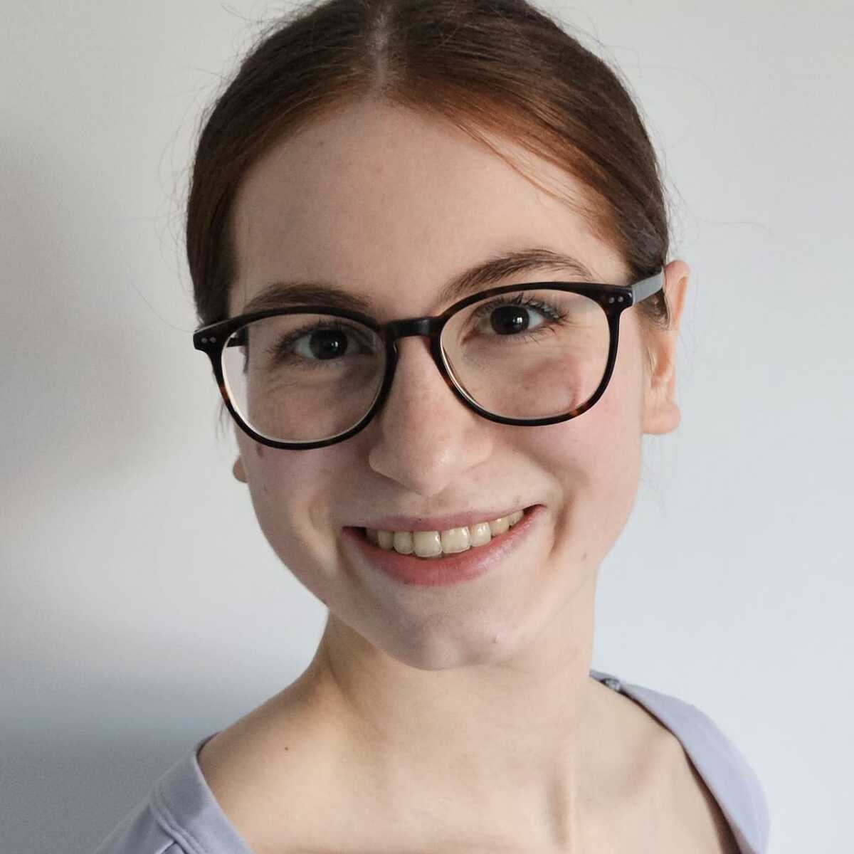 Junge Frau mit Brille l&auml;chelt vor einem hellen Hintergrund und tr&auml;gt ein lila Oberteil.