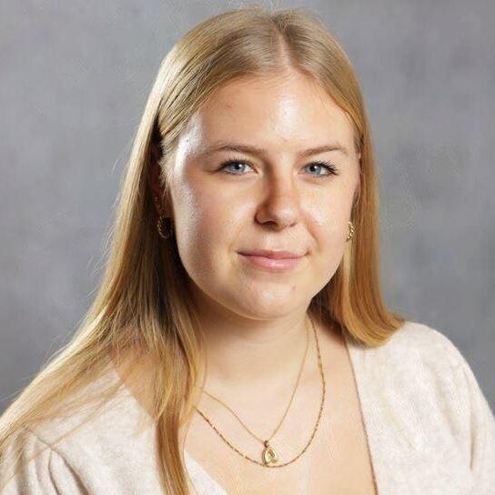 Portr&auml;t einer arbeite jungen Frau mit langen, blonden Haaren, die einen hellen Pullover und eine goldene Kette tr&auml;gt.