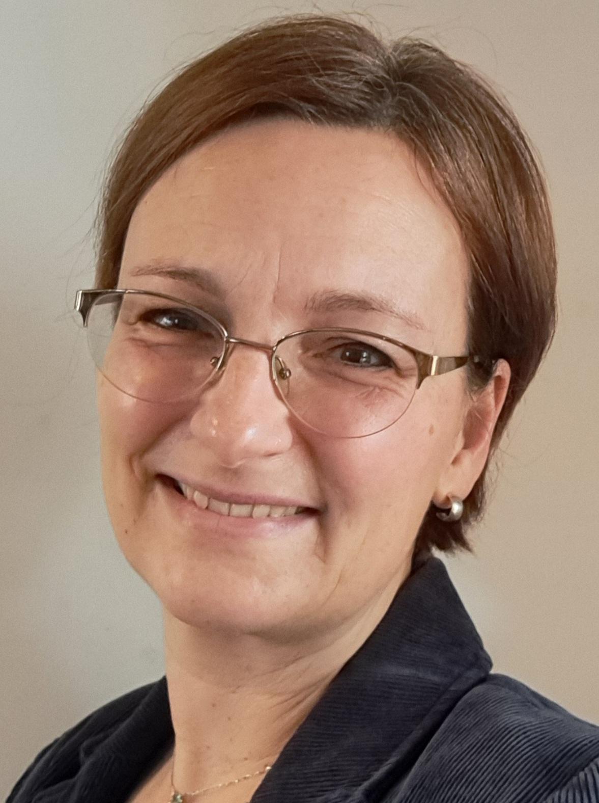 Portr&auml;t einer l&auml;chelnden Frau mit Brille, die einen schwarzen Blazer tr&auml;gt, vor einer neutralen Wand.