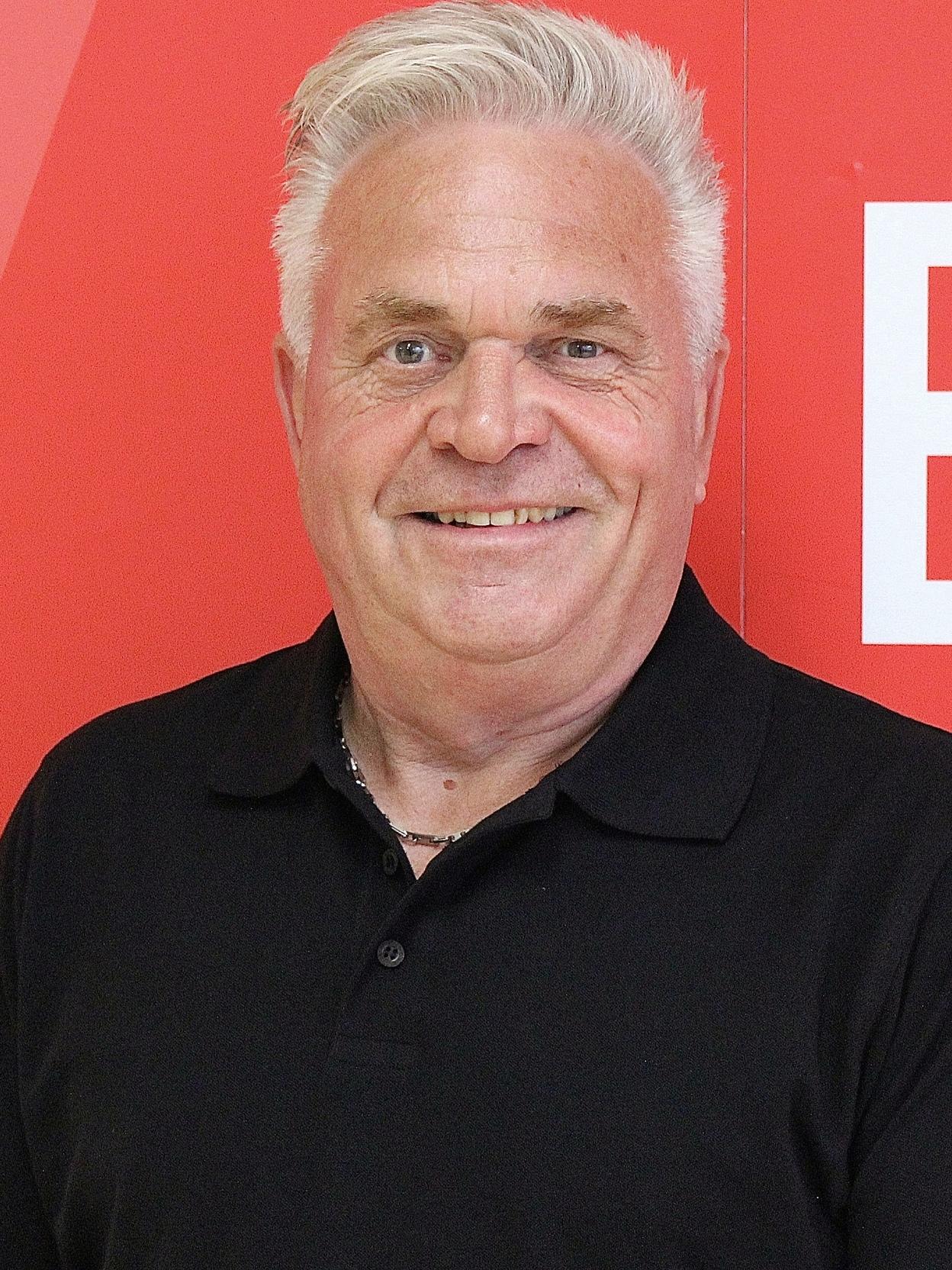 Mann mit kurzem, grau-wei&szlig;en Haar l&auml;chelt vor einer roten Wand mit grafischen Elementen. Tr&auml;gt ein schwarzes Polohemd.