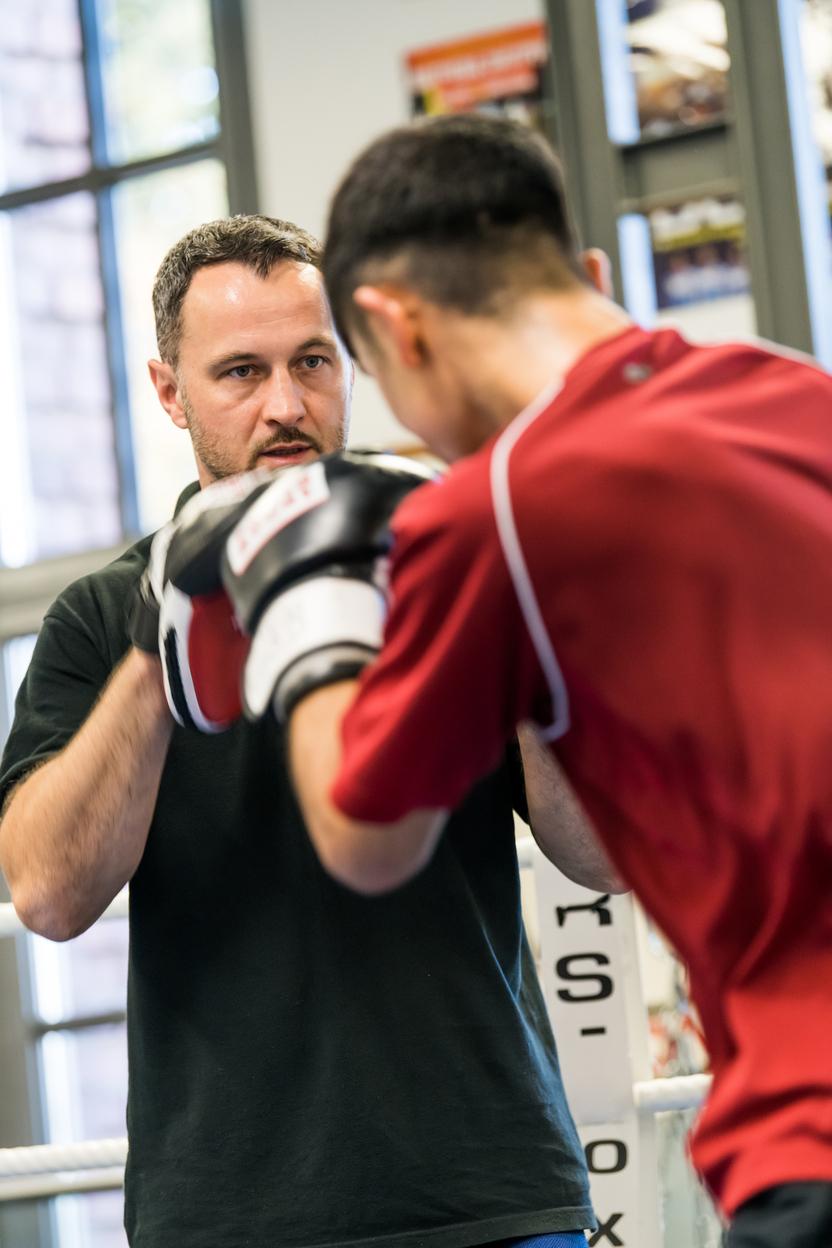 Trainer gibt Anweisungen an einen Boxer in einem Trainingsraum, beide tragen Boxhandschuhe und konzentrieren sich auf Technik.