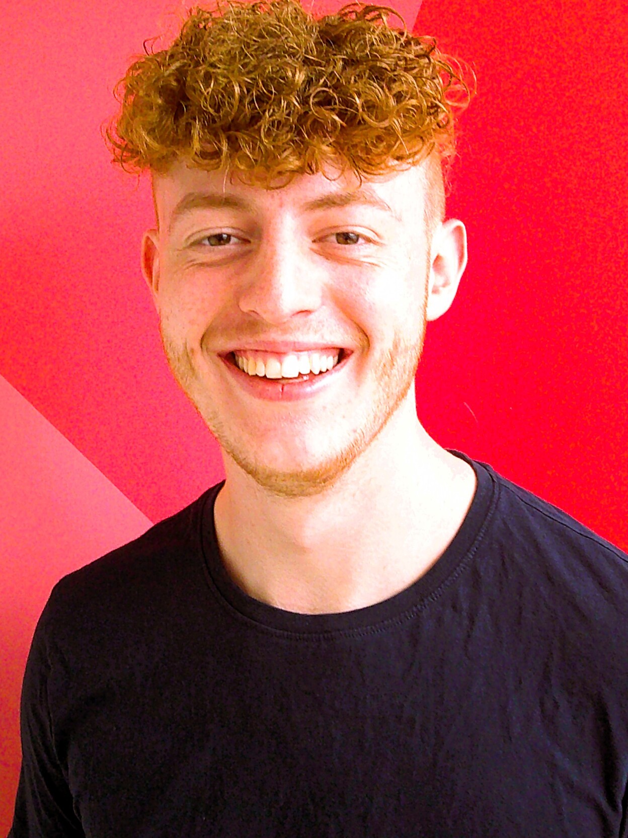 L&auml;chelnder junger Mann mit lockigem, blondem Haar vor einer roten Wand mit geometrischem Muster.