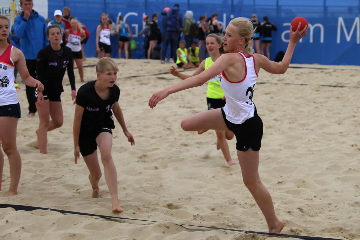 M&auml;dchen im Sand spielt Handball, w&auml;hrend eine Mitspielerin nach dem Ball greift; Zuschauer im Hintergrund.