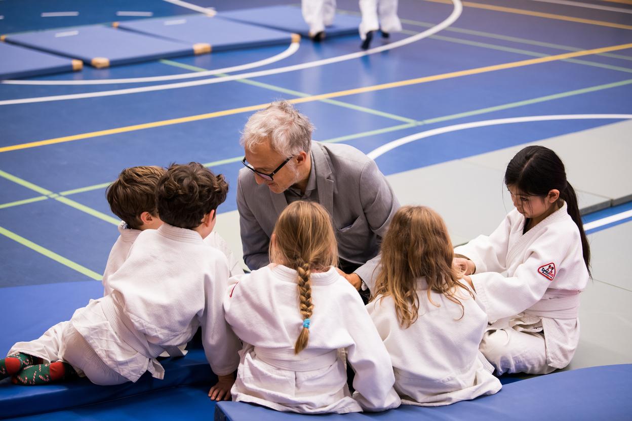 Trainer erkl&auml;rt Judo-Techniken mehreren Kindern in wei&szlig;en Judokis auf einer blauen Judomatte im Trainingsraum.