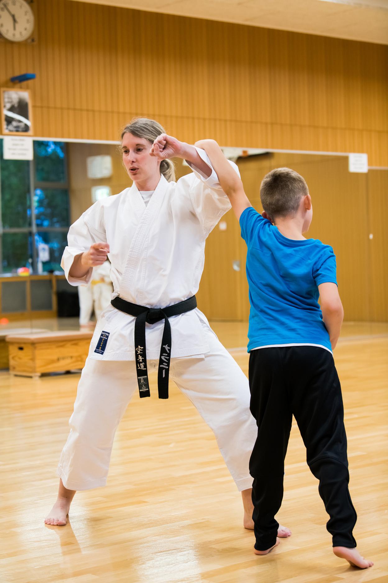 Karateka in wei&szlig;em Gi zeigt einem Kind mit blauer T-Shirt eine Technik in einer Trainingshalle.