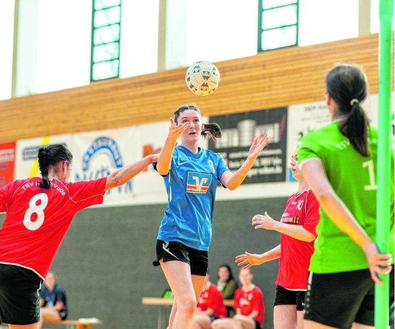 Spielerin in blauer Trikot hebt den Ball zum Wurf, umgeben von Gegnerinnen in roten und gr&uuml;nen Trikots in der Halle.