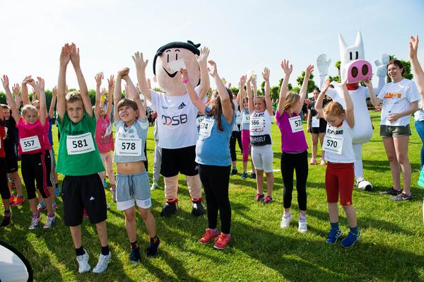 Kinder mit angehobenen Armen stehen zusammen mit Maskottchen auf einer Wiese vor einem sonnigen Himmel.