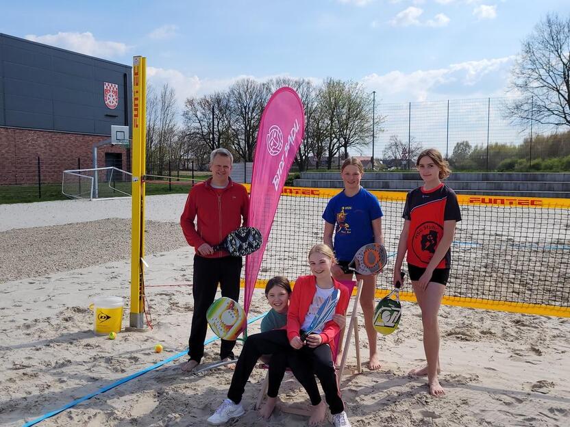 F&uuml;nf Personen stehen und sitzen am Sandplatz mit Netzen, Schl&auml;gern und B&auml;llen, umgeben von B&auml;umen und einem Geb&auml;ude.
