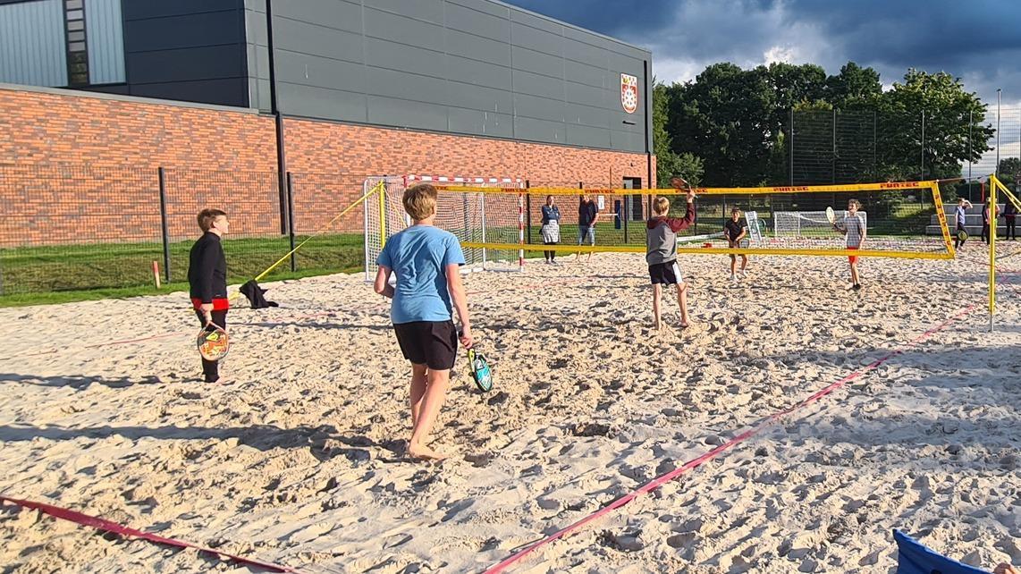 Sechs Kinder spielen Beachvolleyball auf einem Sandplatz mit Netz und angrenzendem Sportgel&auml;nde.