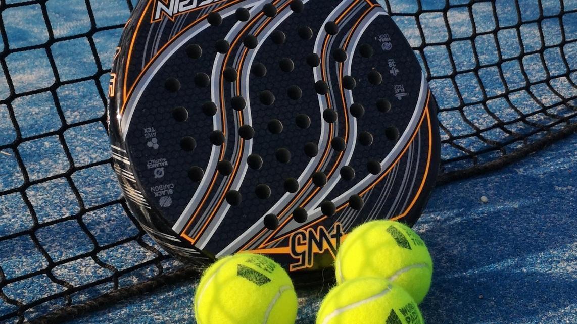 Nahaufnahme eines Padel-Schl&auml;gers und drei gelben B&auml;llen vor einem Netz auf blauem Belag.