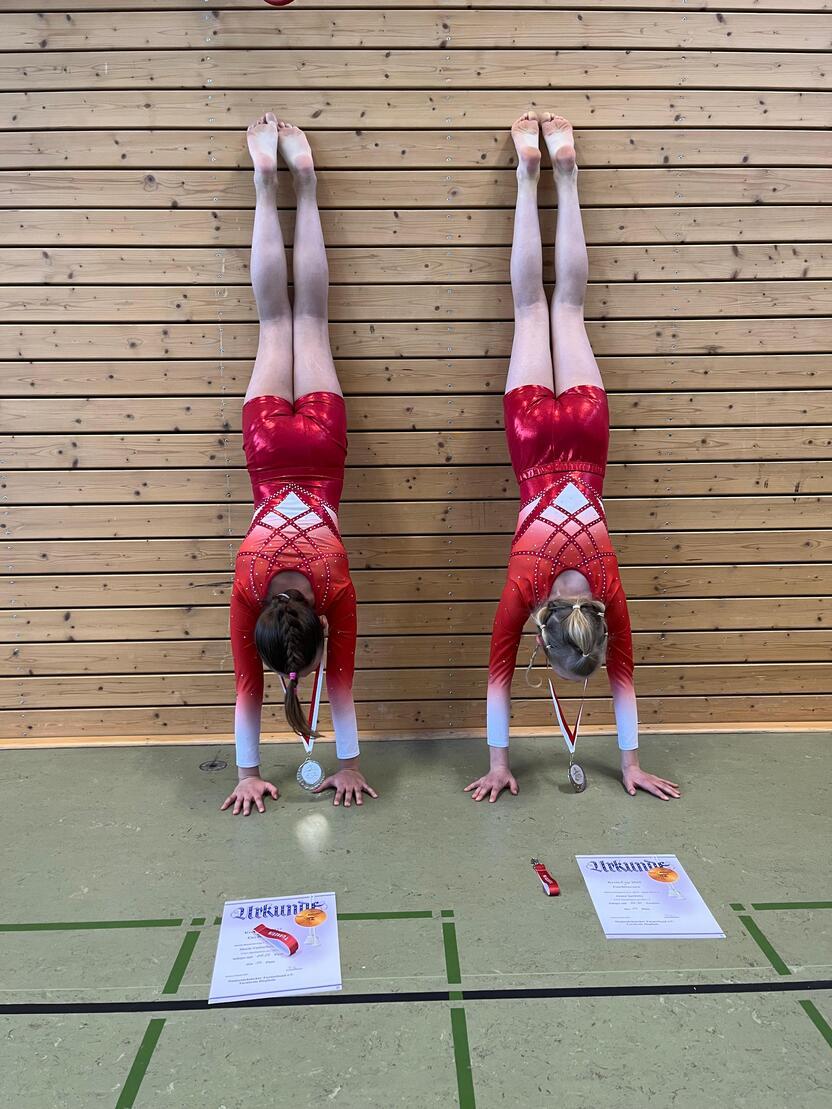 Zwei Turnerinnen in roten Anz&uuml;gen stehen auf H&auml;nden vor einer Holzwand, Medaillen und Urkunden liegen daneben.