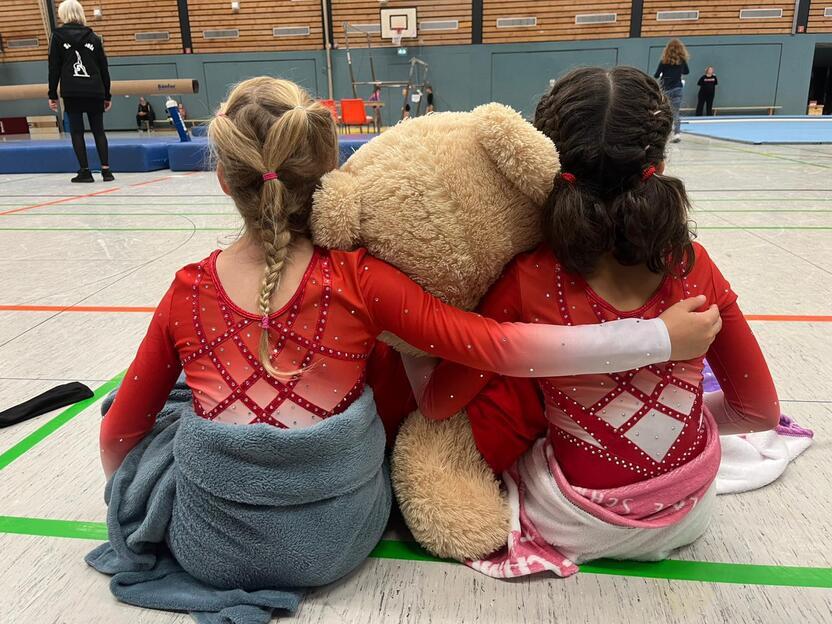 Zwei Kinder in roten Gymnastikanz&uuml;gen sitzen mit einem gro&szlig;en Teddyb&auml;r und genie&szlig;en den Wettkampf.