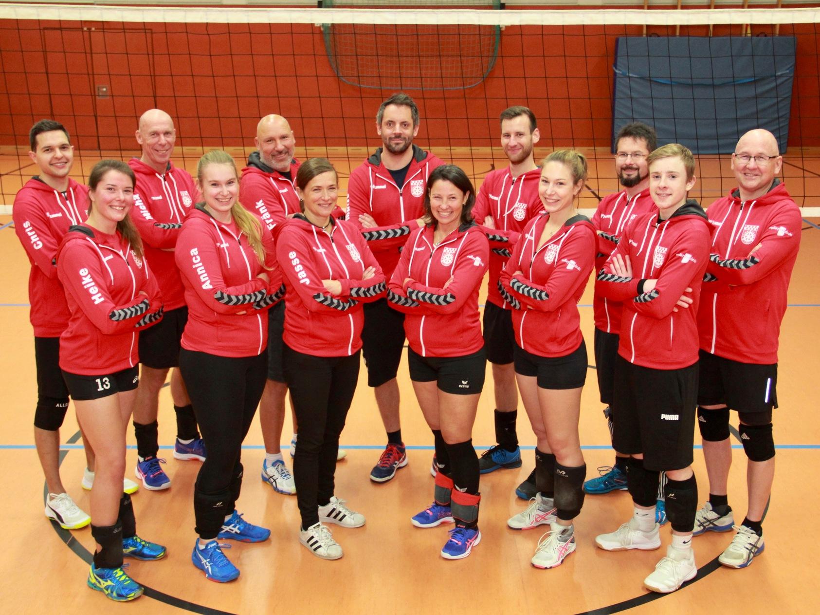 Gruppenfoto von 14 Volleyballspielern in roten Trainingsjacken, aufgeteilt in zwei Reihen vor einem Volleyballnetz.