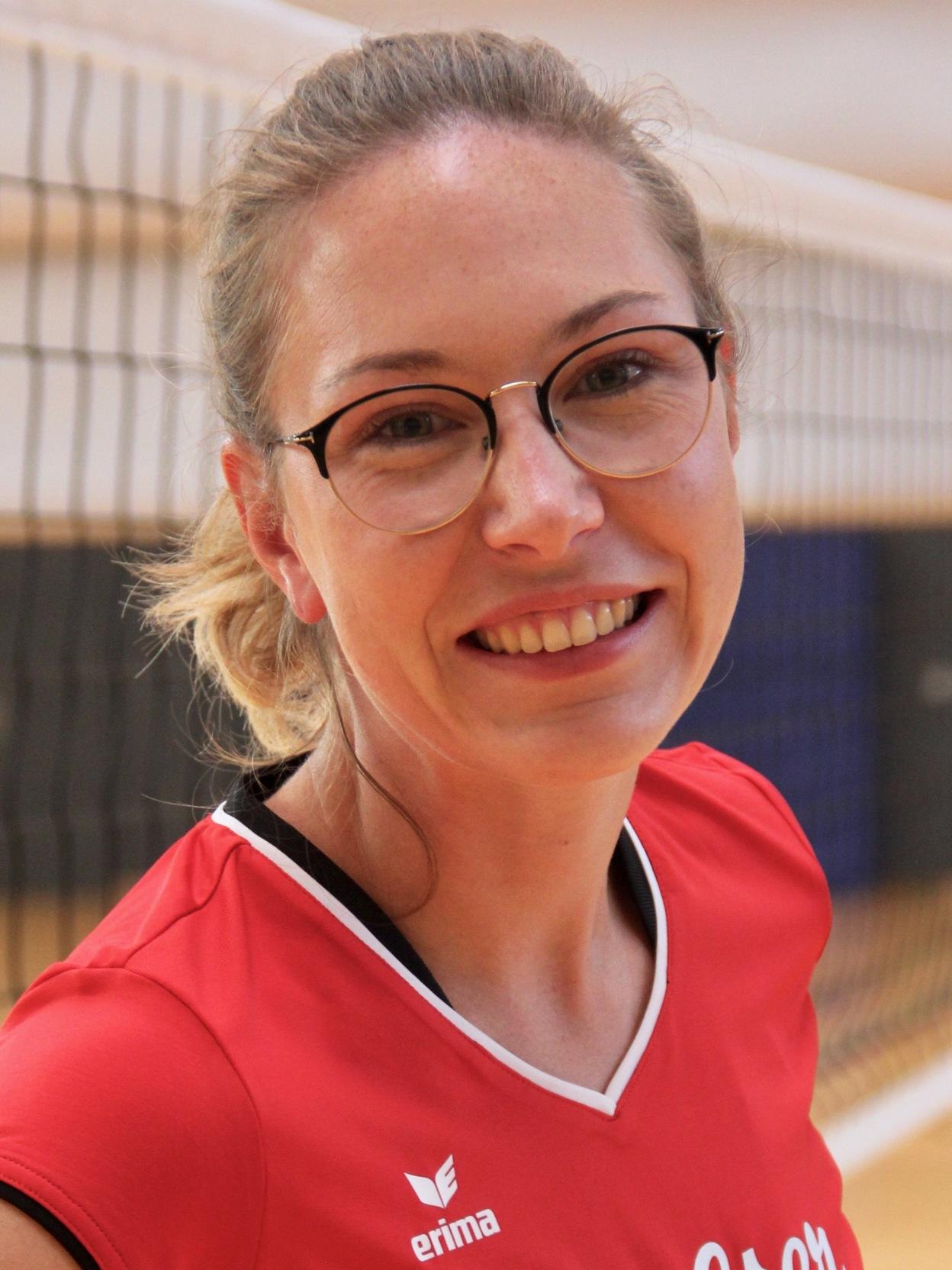 L&auml;chelnde junge Frau in rotem Volleyballtrikot steht vor einem Volleyballnetz in der Halle.