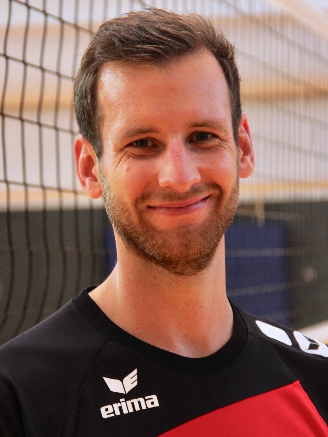 L&auml;chelnder Volleyballspieler in schwarz-rotem Trikot steht vor einem Volleyballnetz in einer Sporthalle.