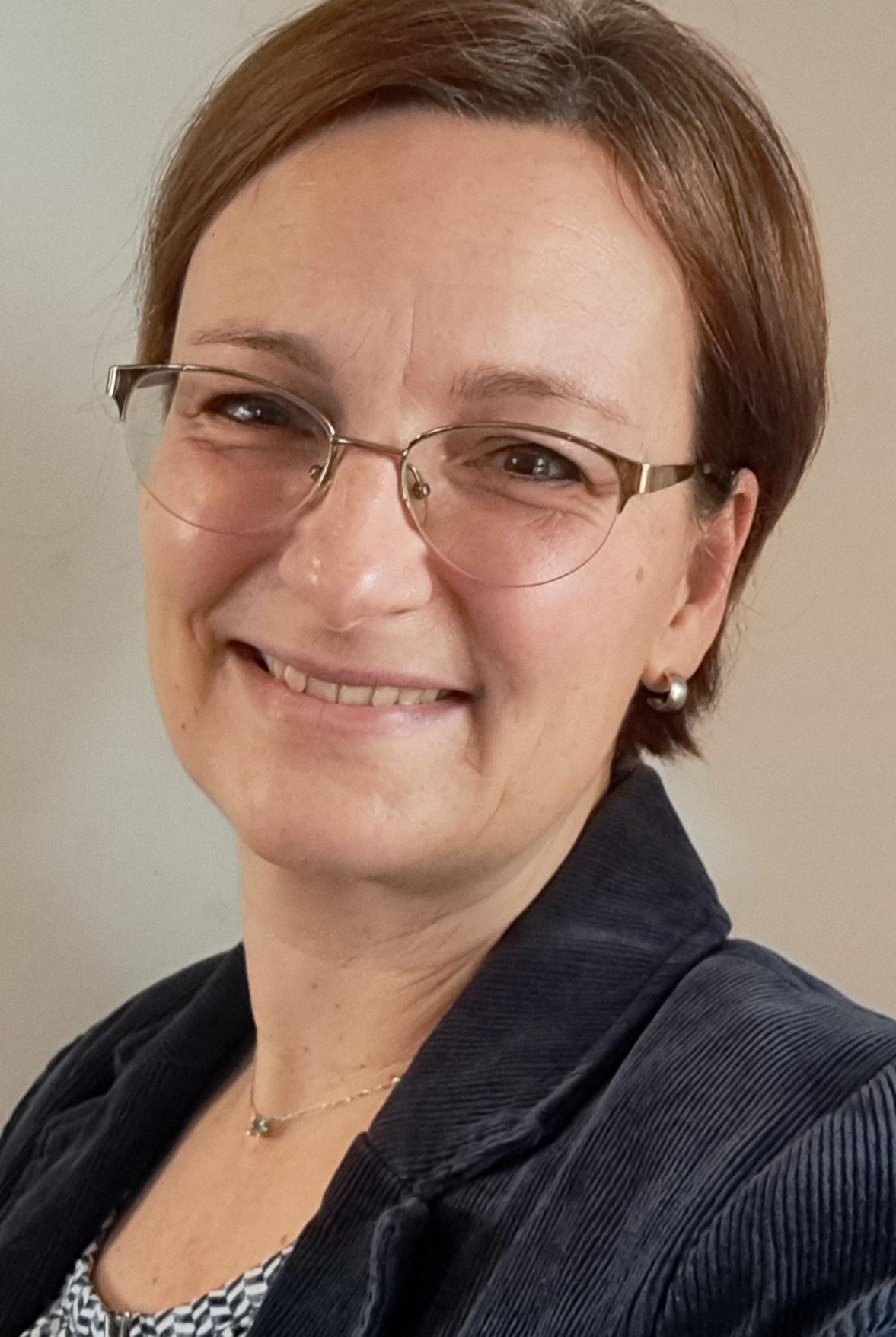 Portr&auml;t einer l&auml;chelnden Frau mit Brille, kurzer brauner Haarfrisur und blauer Jacke, vor neutralem Hintergrund.