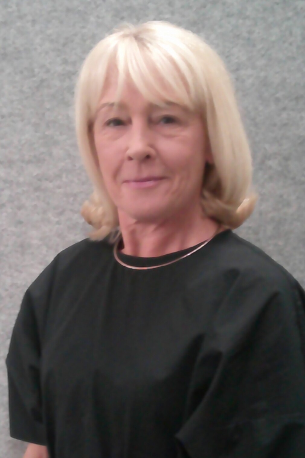 Portr&auml;t einer Frau mit blonden Haare in schwarzer Oberbekleidung vor einem grauen Hintergrund.