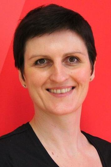 Portr&auml;t einer l&auml;chelnden Frau mit kurzem, dunklem Haar vor rotem Hintergrund. Sie tr&auml;gt ein schwarzes Oberteil.