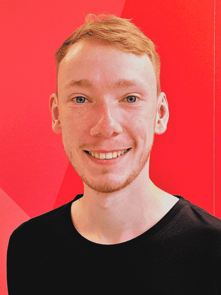 L&auml;chelnder junger Mann mit kurzen, blonden Haaren, tr&auml;gt ein schwarzes Sportshirt vor rotem Hintergrund.