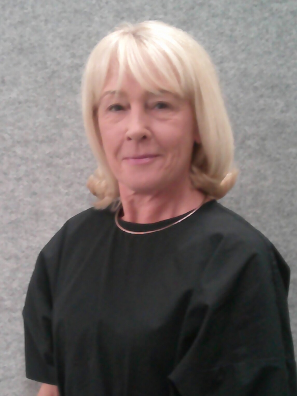 Portr&auml;t einer Frau mit blonden Haare in schwarzer Oberbekleidung vor einem grauen Hintergrund.