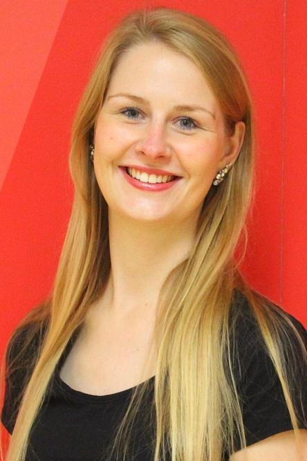 L&auml;chelnde Frau mit langen, blonden Haaren und Ohrringen vor roter Wand. Sie tr&auml;gt ein schwarzes Oberteil.