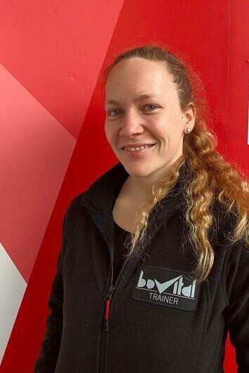 L&auml;chelnde Frau mit langen, lockigen Haaren, tr&auml;gt einen schwarzen Trainer-Pullover vor rotem Hintergrund.