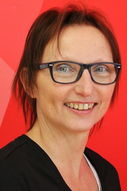 Portr&auml;t einer l&auml;chelnden Frau mit Brille vor rotem Hintergrund. Sie tr&auml;gt ein schwarzes T-Shirt und hat kurze, braune Haare.