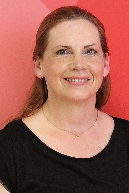 Portr&auml;t einer l&auml;chelnden Frau mit langen, roten Haaren vor einem roten Hintergrund. Sie tr&auml;gt ein schwarzes Oberteil.