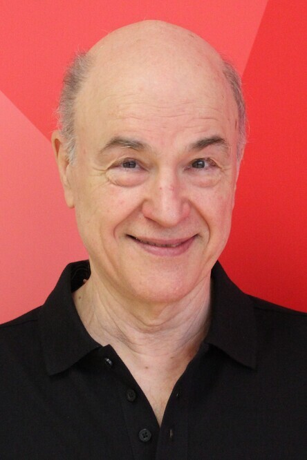 Portr&auml;t eines l&auml;chelnden Mannes mit Glatze und schwarzem Polohemd vor einem roten geometrischen Hintergrund.