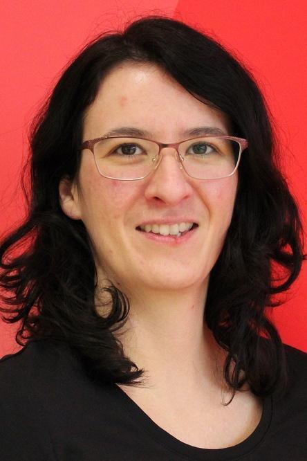 Portr&auml;t einer l&auml;chelnden Frau mit dunklen, lockigen Haaren auf rotem Hintergrund. Sie tr&auml;gt eine schwarze Bluse und Brille.