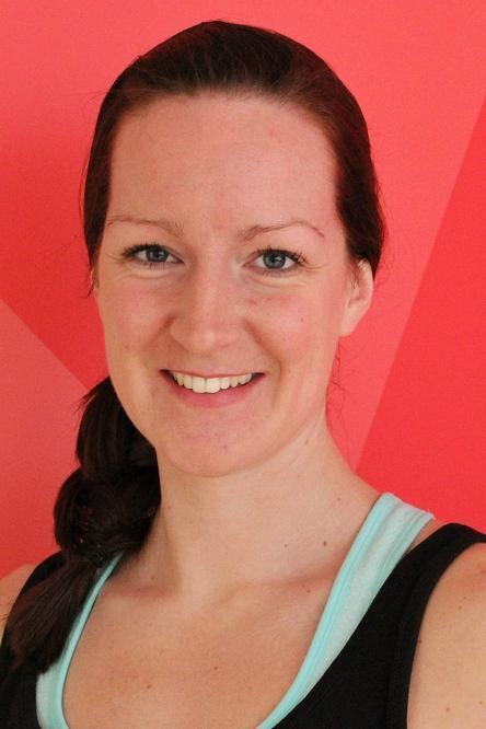 L&auml;chelnde Frau mit langen, braunen Haaren vor einem pinken, geometrischen Hintergrund. Sie tr&auml;gt ein schwarzes Oberteil.