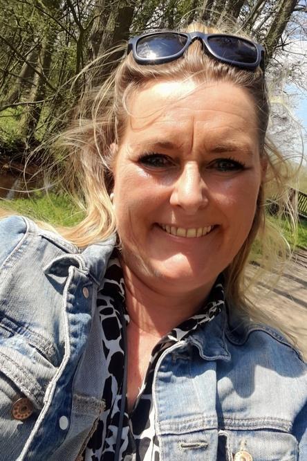 Frau mit blonden Haaren und Sonnenbrille l&auml;chelt vor einer gr&uuml;nen, nat&uuml;rlichen Umgebung. Sie tr&auml;gt eine Jeansjacke und ein gemustertes Oberteil.