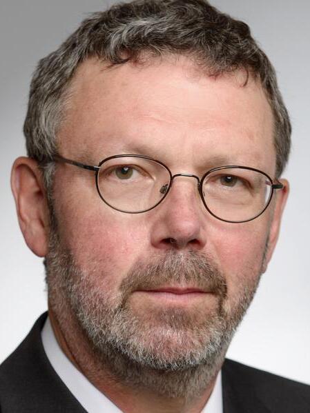 Portr&auml;t eines Mannes mit Brille, grauem Haar und Bart, gekleidet in einem dunklen Anzug mit kariertem Schlips.