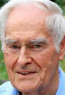 &Auml;lterer Mann mit grauen Haaren und freundlichem Gesichtsausdruck, im Hintergrund unscharfe gr&uuml;ne Pflanzen.