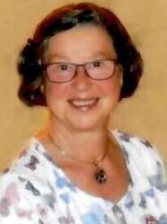 Eine l&auml;chelnde Frau mit Brille und schulterlangen, lockigen Haaren tr&auml;gt ein bunten Oberteil und Halskette.