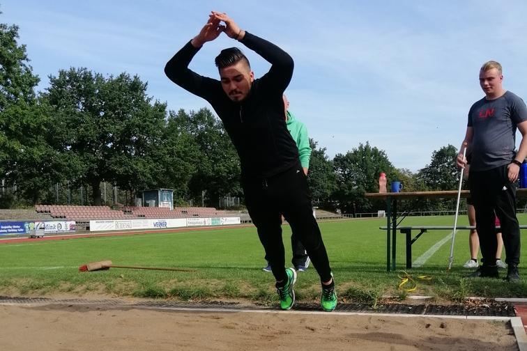 Sportler springt in den Sandgrube eines Athletikfeldes, w&auml;hrend zwei Personen im Hintergrund zuschauen.