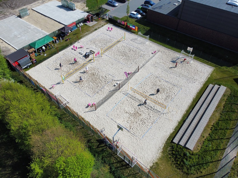 Luftaufnahme eines Sandfelds mit zwei Volleyballfeldern und Spielern, umgeben von Gras und einem Geb&auml;ude im Hintergrund.